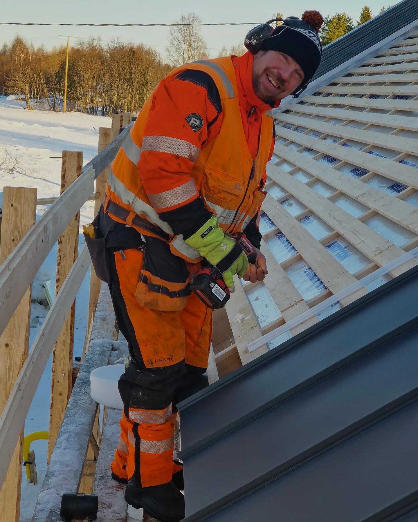Rakennustyöntekijä oranssissa työtakissa ja pipossa katolla, pitäen akkuporakonetta kädessään, hymyillen kameraan, aurinkoisessa säässä.